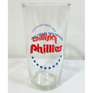 Pode incluir: Um copo de vidro transparente com o texto vermelho Phillies e o texto azul The Star Spangled Banner arqueado acima. Abaixo do logótipo vermelho Phillies, existe uma fila curva de doze estrelas azuis impressas na superfície do vidro.