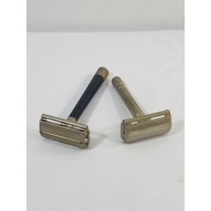 Puede incluir: Dos maquinillas de afeitar de seguridad vintage. Una tiene un mango negro texturizado y la otra un mango de metal liso. Ambas maquinillas tienen un cabezal y un protector de cuchilla plateados.