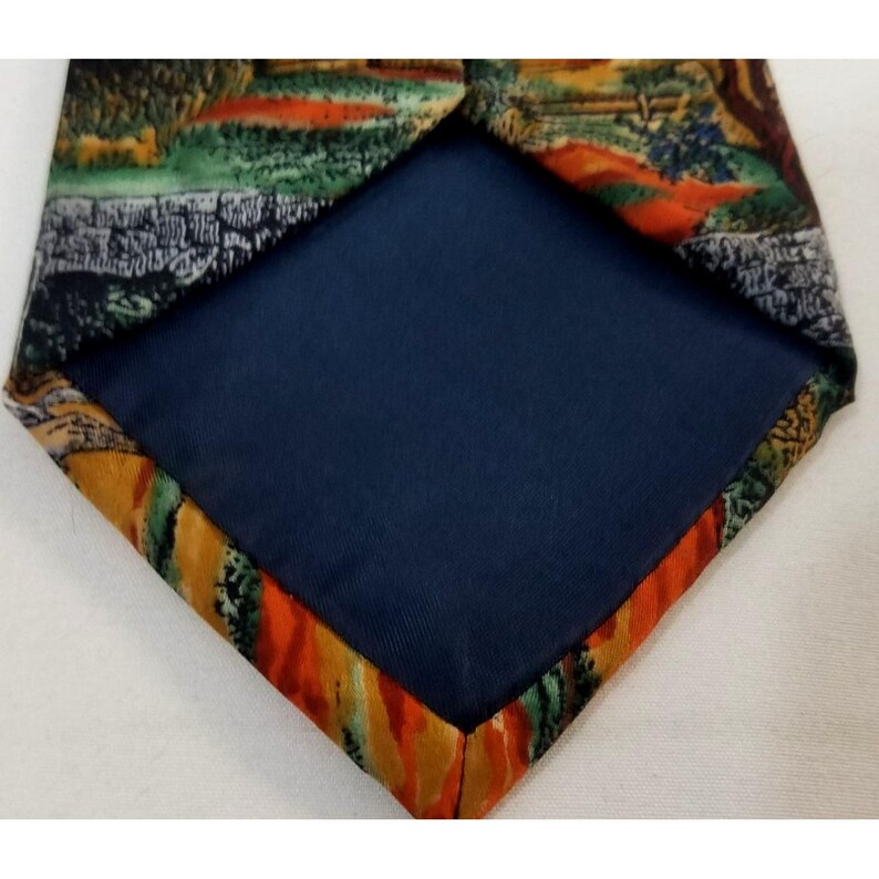 May include: A necktie with a landscape design on the front and a solid navy blue backing. The landscape features green, orange, and brown hues. The tie's edges have a colorful, abstract pattern.