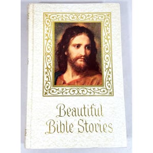 May include: A white hardcover book titled Beautiful Bible Stories in gold lettering. The cover features a central rectangular portrait of a bearded man with long brown hair, enclosed in an ornate gold filigree frame against a textured white background.