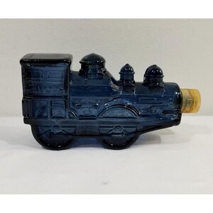 May include: A dark blue glass train-shaped bottle with a gold-colored stopper. The bottle has embossed details and the word "AVON" is visible on the side. The train has wheels and a detailed engine design.