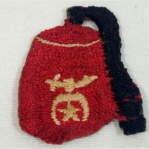 Op de afbeelding: Een rood geborduurd embleem met een zwarte handgreep. Het embleem heeft een gouden embleem met een ster en andere symbolen. Het embleem is waarschijnlijk een decoratief item of embleem.