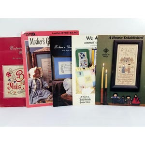 May include: A collection of vintage cross-stitch pattern books. The covers feature images of dolls, framed samplers, and floral designs. Titles include "Mother's G", "A House Established", and "Jeremiah Junction".