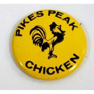 May include: Yellow button with black text and a black rooster illustration. The text reads "Pikes Peak Chicken".