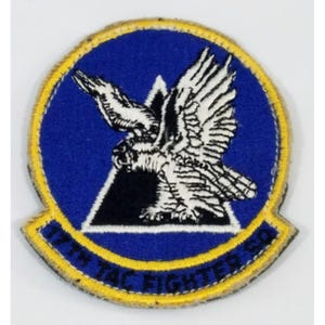 May include: A circular patch with a blue background, featuring a white and black eagle design. The patch is edged with yellow and has the text "17th TAC FIGHTER SQ". The design includes a black triangle.