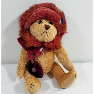 Pode incluir: Um urso de pelúcia com pelo castanho claro, usando um chapéu e cachecol bordô. O chapéu tem uma aparência fofa e texturizada com tons de laranja e vermelho. O urso segura uma pequena bolsa vermelha escura com um detalhe de rosa.