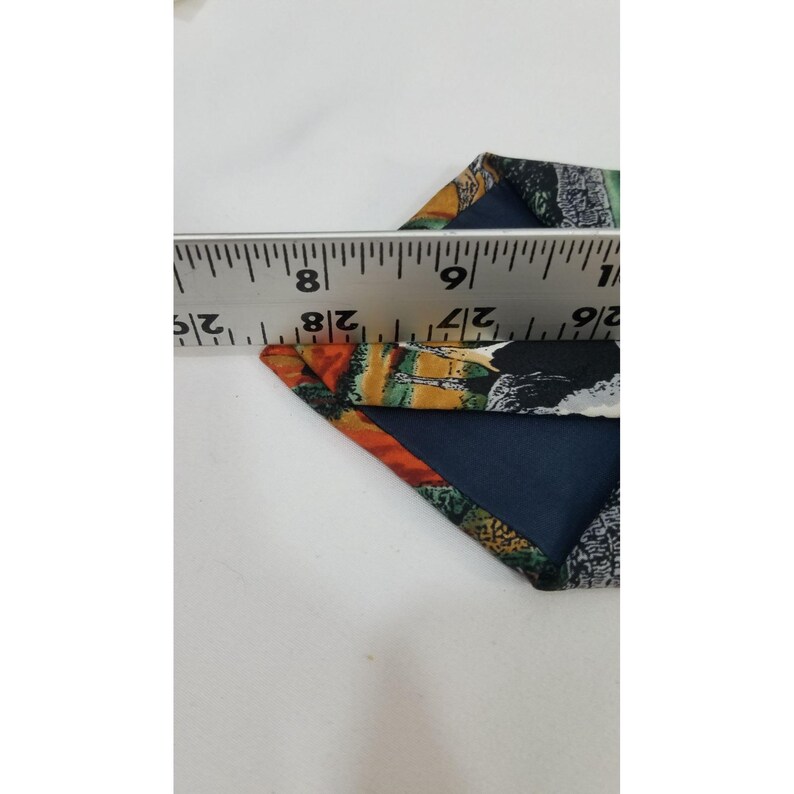 May include: A patterned necktie with a dark blue base, featuring orange, green, and black abstract designs. The tie is measured with a ruler, showing a width of approximately 2.5 inches.