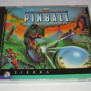 Puede incluir: Una caja de CD para el juego "Ultra 3D Pinball: The Lost Continent" de Sierra. La portada presenta un dinosaurio verde con un orbe azul brillante en el fondo.