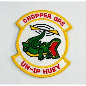 May include: Embroidered patch with a white background, yellow border, and the words "CHOPPER OPS" and "UH-1P HUEY" in red. The patch features a green alligator with an open red mouth and a yellow helicopter.