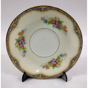 May include: A white porcelain saucer with a gold and black rim and a floral design. The saucer has a scalloped edge and is decorated with a delicate floral pattern in shades of pink, yellow, and blue.