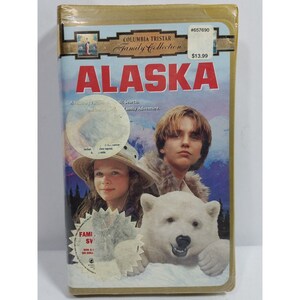 May include: A VHS tape cover for the movie "Alaska" featuring a young boy and girl with a polar bear. The cover is gold with a blue and white background. The text "#657690", "Columbia Tristar Family Collection", "$13.99", and "Alaska" is visible on the cover.