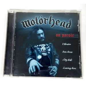 Könnte beinhalten: Ein Motörhead-CD-Album mit dem Titel "on parole" mit einem Bandmitglied in einer Lederjacke. Das Albumcover hat einen dunkelblauen Hintergrund mit dem Namen der Band in einer stilisierten Schriftart. Zu den Titeln gehören "Vibrator" und "Iron Horse".