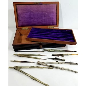 May include: A wooden storage box with a purple velvet-lined interior and a removable tray, accompanied by a collection of vintage metal drafting compasses and technical drawing instruments arranged on a surface in front of the open case.