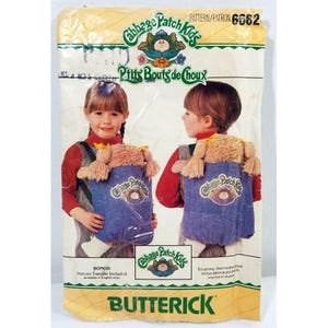 May include: A vintage Butterick sewing pattern for Cabbage Patch Kids dolls. The pattern packet shows two children wearing doll carriers. The packet has the text "Cabbage Patch Kids" and "Prits Bouts de Choux".
