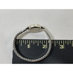 May include: A silver wristwatch with a square face and a silver metal band. The watch is about 1.5 inches in diameter and is shown on a ruler.