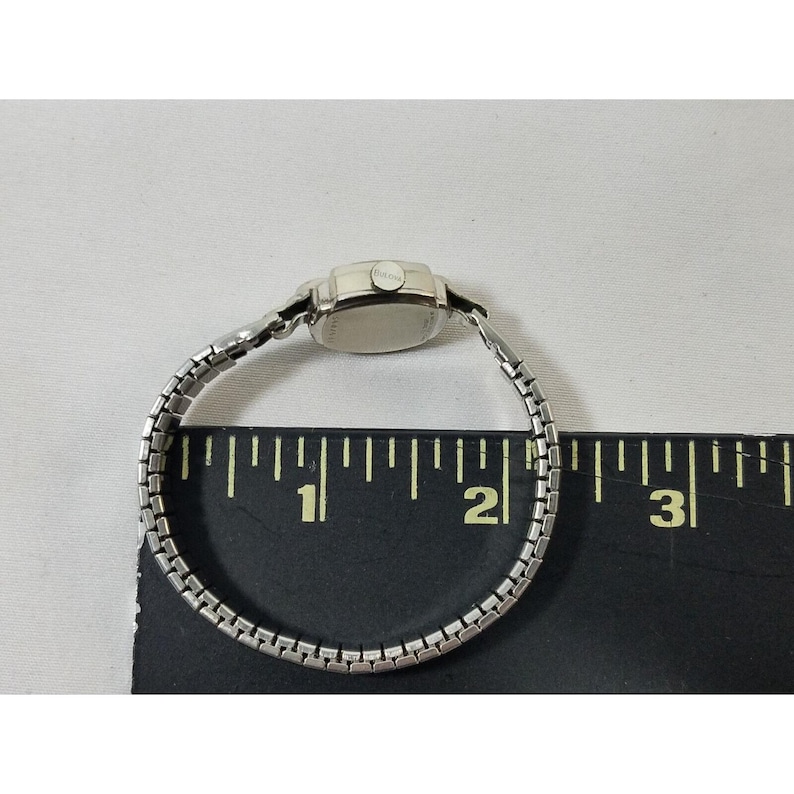 May include: A silver wristwatch with a square face and a silver metal band. The watch is about 1.5 inches in diameter and is shown on a ruler.