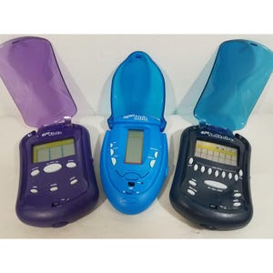 May include: Three handheld electronic games with flip-top lids. The left is purple with Slots text, the center is blue with Tetris text, and the right is dark blue with Solitaire text. Each device features a monochrome LCD screen and control buttons.