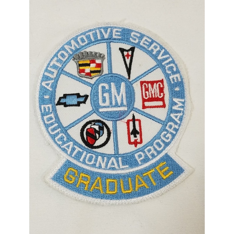 Pode incluir: Um remendo bordado azul e branco com o texto "Automotive Service Educational Program Graduate" e os logotipos de v&aacute;rias marcas da General Motors, incluindo Chevrolet, Cadillac, GMC e Pontiac.