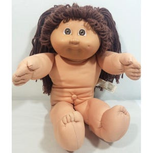 Puede incluir: Una muñeca de cuerpo blando con cara redonda de tela color melocotón, pelo de lana marrón y ojos marrones. La muñeca tiene una nariz pequeña, un hoyuelo en la mejilla izquierda y un ombligo. Brazos y piernas extendidos, sin ropa, etiqueta de tela visible.