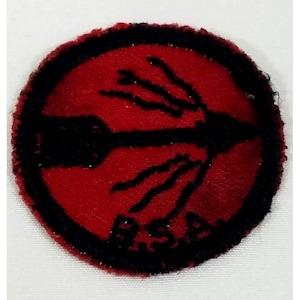May include: A round, red patch with a black border and a black bow and arrow design. The letters "B.S.A." are visible in black at the bottom of the patch. The patch has a textured, felt-like appearance.