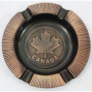 Może przedstawiać: Popielniczka w kolorze czarnym i miedzianym z wypukłym wzorem liścia klonu i napisem "CANADA" wokół.