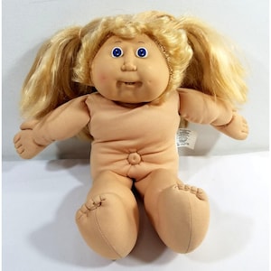 May include: A vintage Cabbage Patch Kid doll with blonde hair styled in pigtails. The doll has blue eyes, a peach-colored face, and a soft, tan body. It is sitting with its arms outstretched and its legs extended.