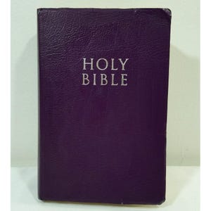 May include: A purple hardcover book with the words "HOLY BIBLE" in silver lettering. The book has a textured cover and is standing upright. The edges of the book show slight wear.