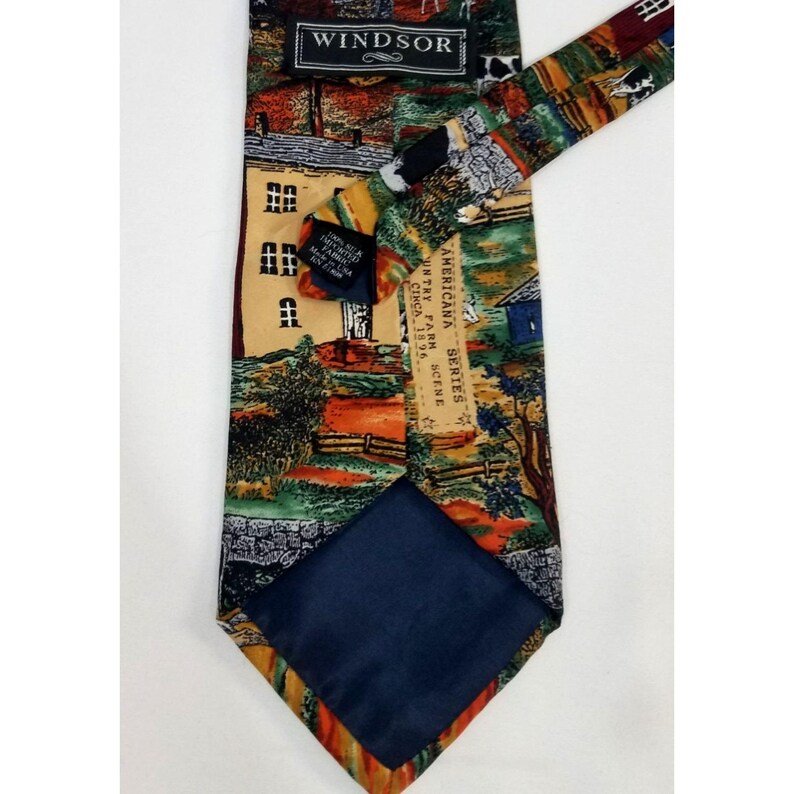 May include: A Windsor brand necktie with a colorful Americana scene print. The tie features a farm scene with a house, trees, and a barn. The tie is made of a silky material and has a navy blue backing. The text "Windsor" is visible.