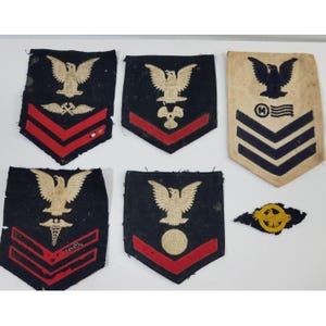 Puede incluir: Colección de parches militares vintage. Los parches presentan un emblema de águila, galones y varios símbolos. Los parches son principalmente azul marino con detalles en rojo y dorado. Un parche es de color beige con detalles en negro y azul.