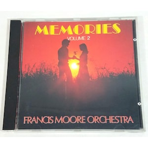 May include: A compact disc case featuring the album title MEMORIES VOLUME 2 in yellow text at the top. The cover art displays a silhouette of two figures standing in a field against a bright orange sunset. The artist name FRANCIS MOORE ORCHESTRA is printed in red at the bottom.