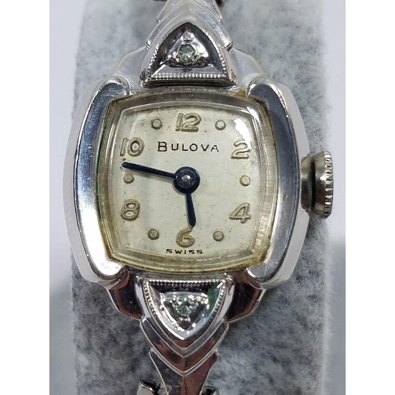 May include: A silver Bulova wristwatch with a square face and a diamond-encrusted band. The watch face has black hands and Roman numeral hour markers. The watch face also has the word "Bulova" and "Swiss" printed on it.