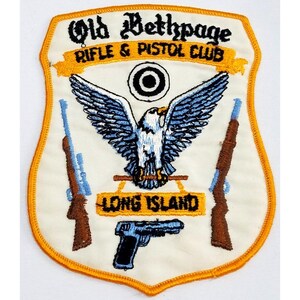 May include: A shield-shaped patch with the text "Old Bethpage Rifle & Pistol Club" and "Long Island." The patch features an eagle with outstretched wings, rifles, and a pistol. The border is gold.