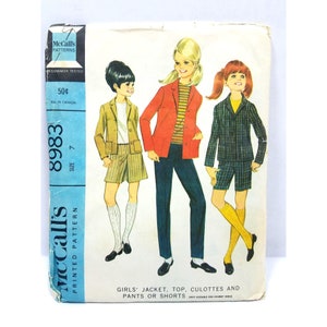 May include: A vintage McCall's sewing pattern for girls' clothing. The pattern is for a jacket, top, culottes, and pants or shorts. The pattern number is 8983 and the size is 7. The pattern is printed on a blue and white background with a picture of three girls wearing the different garments.