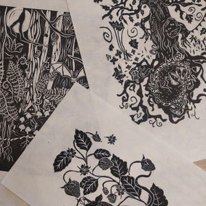 Może przedstawiać: Kolekcja czarno-białych linorytów na papierze. Grafiki przedstawiają scenę leśną z nietoperzem, dużym drzewem z lisem i wiewiórką z truskawkami. Dzieła sztuki są ułożone na jasnej powierzchni.