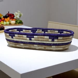 Puede incluir: Una cesta ovalada tejida con un patrón de rayas azules, beige y blancas. La cesta está hecha de fibras naturales y está sobre una superficie blanca. La cesta está vacía y parece ser un artículo decorativo.