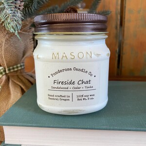 May include: A white Mason jar candle with a brown lid. The label reads "Fireside Chat" with scent notes of sandalwood, cedar, and tonka. It is 8 oz. and hand crafted in Central Oregon.