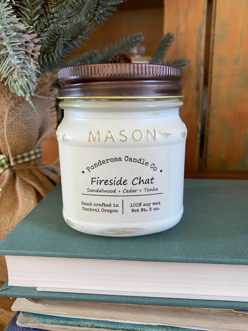 May include: A white Mason jar candle with a brown lid. The label reads "Fireside Chat" with scent notes of sandalwood, cedar, and tonka. It is 8 oz. and hand crafted in Central Oregon.