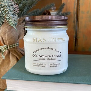 May include: A white Mason jar candle with a brown lid. The label reads "Old Growth Forest" with "Cypress + Bayberry" and "100% soy wax Net Wt. 8 oz." The candle is on a stack of books.