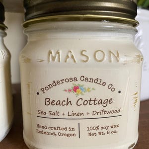 May include: A small, off-white Mason jar candle with a bronze lid. The label reads "Beach Cottage" with "Sea Salt + Linen + Driftwood" scent. It is hand-crafted in Redmond, Oregon, and contains 8 oz of 100% soy wax.