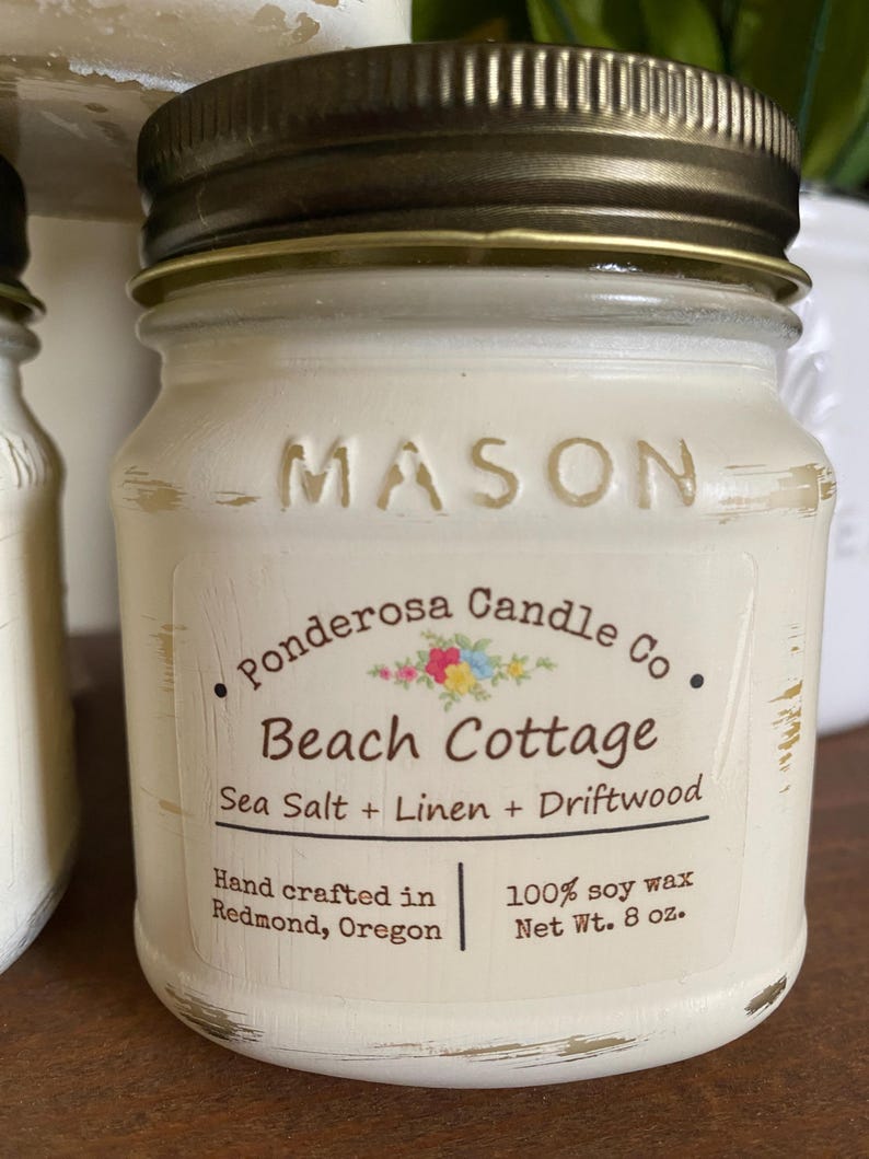 May include: A small, off-white Mason jar candle with a bronze lid. The label reads "Beach Cottage" with "Sea Salt + Linen + Driftwood" scent. It is hand-crafted in Redmond, Oregon, and contains 8 oz of 100% soy wax.