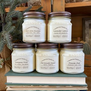 May include: Six mason jar candles with brown lids are displayed on a stack of books. The labels read "Ponderosa Candle Co." with scents like "Mint White Chocolate" and "Lavender Lemon." Each candle is 8 oz.