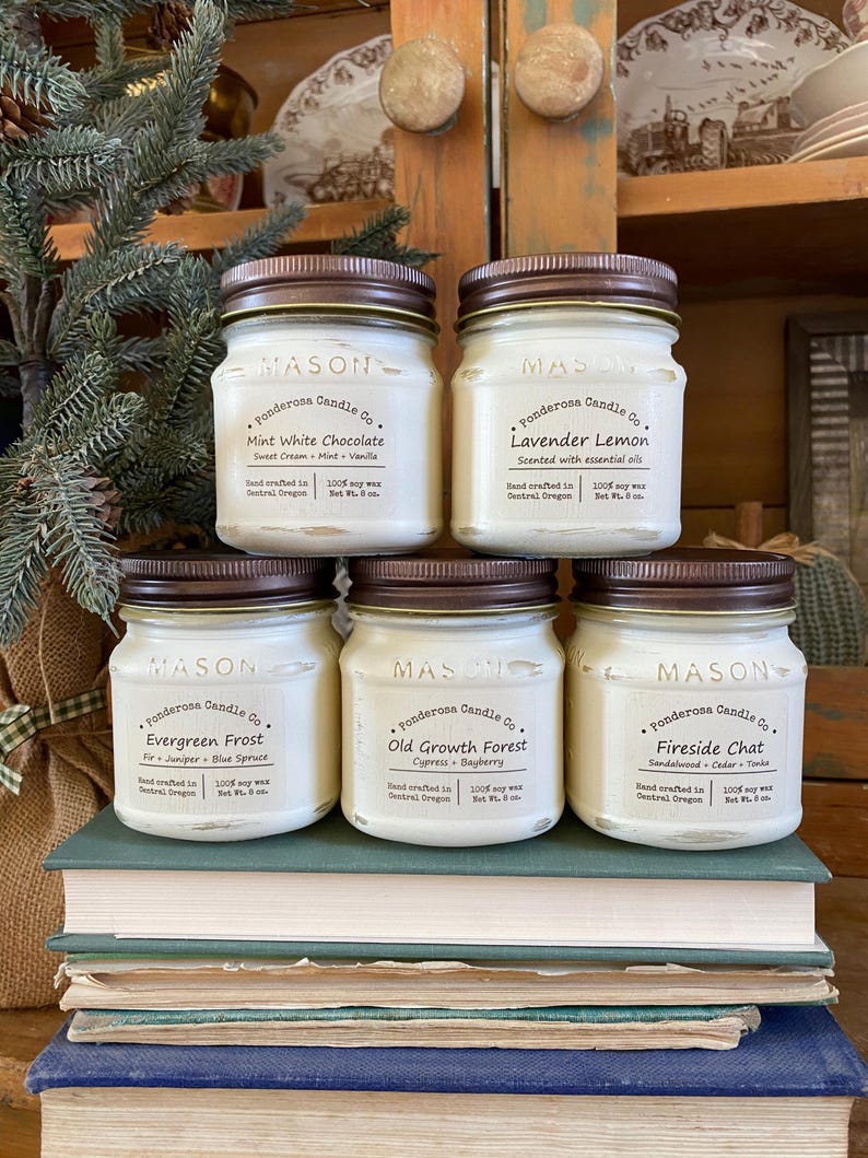 May include: Six mason jar candles with brown lids are displayed on a stack of books. The labels read "Ponderosa Candle Co." with scents like "Mint White Chocolate" and "Lavender Lemon." Each candle is 8 oz.