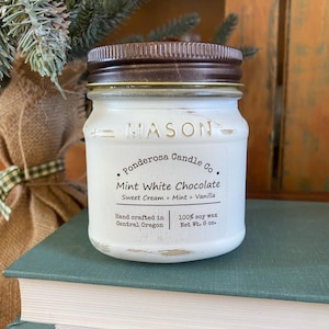May include: A white Mason jar candle with a brown lid. The label reads "Mint White Chocolate" with "Sweet Cream + Mint + Vanilla" and "100% soy wax Net Wt. 8 oz.". The candle is on a stack of books.