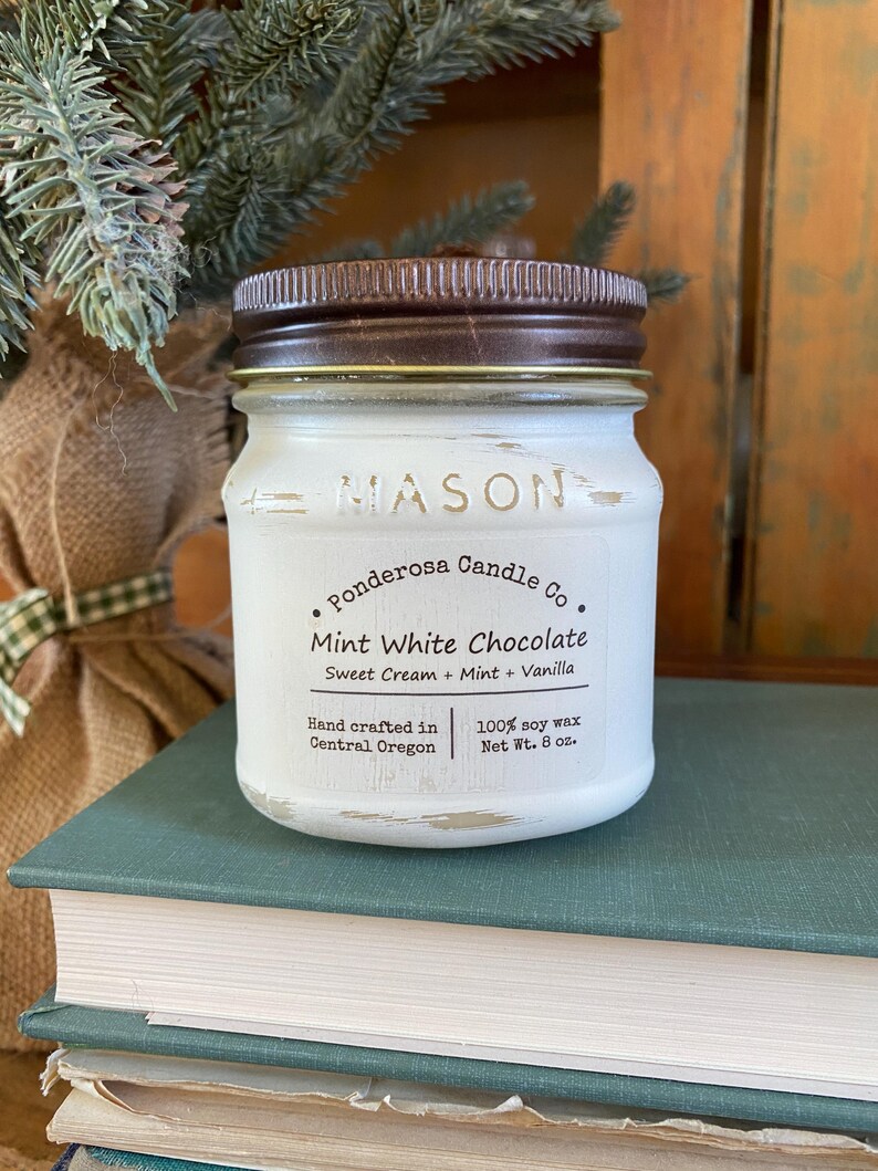 May include: A white Mason jar candle with a brown lid. The label reads "Mint White Chocolate" with "Sweet Cream + Mint + Vanilla" and "100% soy wax Net Wt. 8 oz.". The candle is on a stack of books.