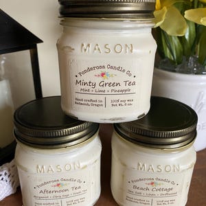 May include: Three white Mason jar candles with black lids are stacked. The labels read "Minty Green Tea", "Afternoon Tea", and "Beach Cottage". Each candle is from Ponderosa Candle Co. and made with 100% soy wax. The jars are 8 oz.
