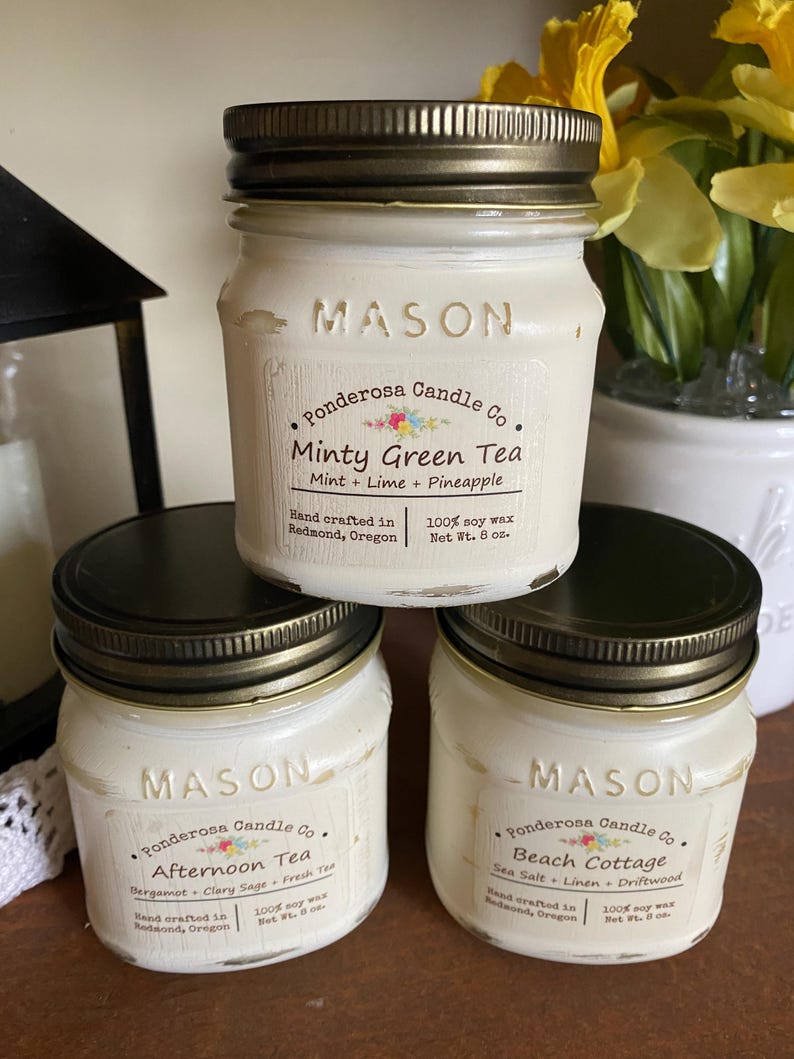 May include: Three white Mason jar candles with black lids are stacked. The labels read "Minty Green Tea", "Afternoon Tea", and "Beach Cottage". Each candle is from Ponderosa Candle Co. and made with 100% soy wax. The jars are 8 oz.