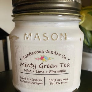 May include: A white Mason jar candle with a bronze lid. The label reads "Minty Green Tea" with mint, lime, and pineapple scents. The candle is hand-crafted in Redmond, Oregon, and made with 100% soy wax, weighing 8 oz.