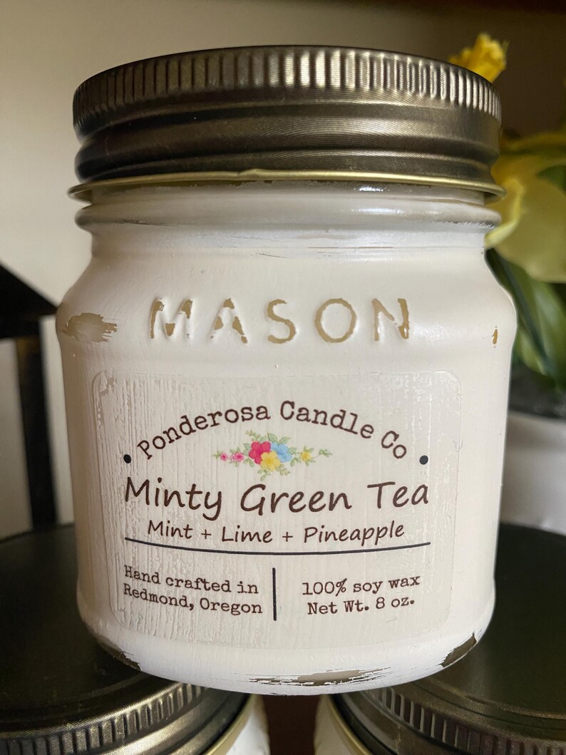 May include: A white Mason jar candle with a bronze lid. The label reads "Minty Green Tea" with mint, lime, and pineapple scents. The candle is hand-crafted in Redmond, Oregon, and made with 100% soy wax, weighing 8 oz.
