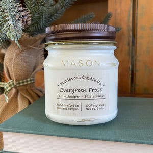 May include: A white Mason jar candle with a brown lid, labeled "Evergreen Frost." The label includes "Fir + Juniper + Blue Spruce" and "100% soy wax, Net Wt. 8 oz." The candle sits on a stack of books, with greenery in the background.