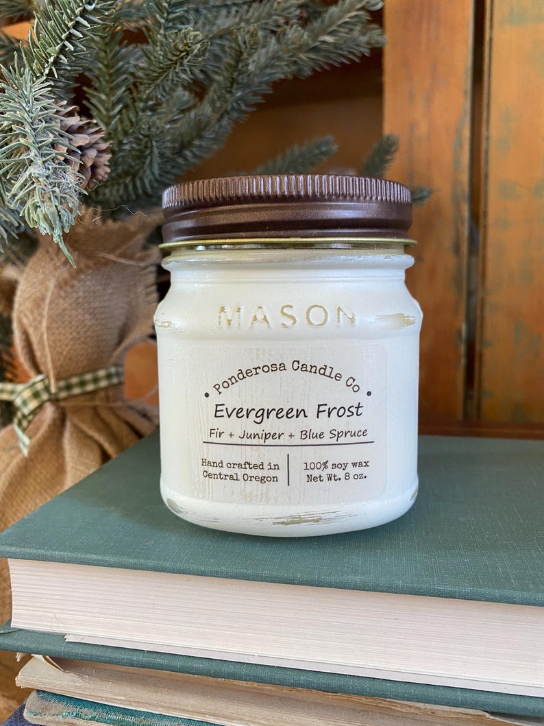 May include: A white Mason jar candle with a brown lid, labeled "Evergreen Frost." The label includes "Fir + Juniper + Blue Spruce" and "100% soy wax, Net Wt. 8 oz." The candle sits on a stack of books, with greenery in the background.
